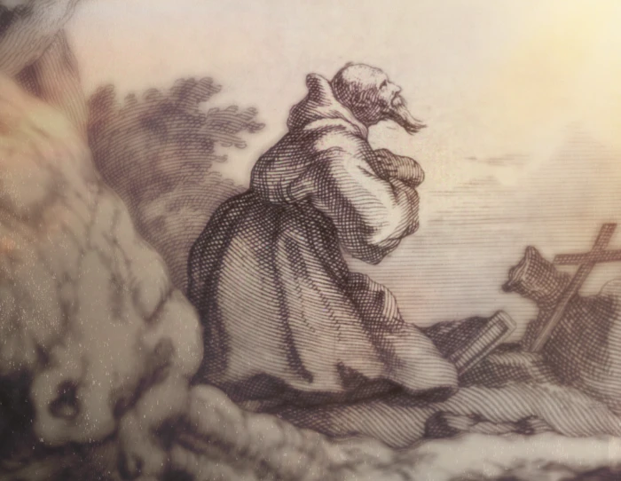 A hermit kneels at the crest of a small hill, praying while looking upward into the sky. A tree precariously stands behind the man and a cross and prayer book lay before the kneeling person.